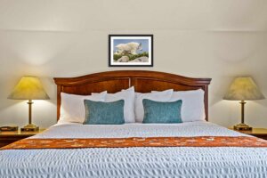 Spacious and comfortable bed in the small suite with blue pillows, matching table lamps, an orange accent blanket, and artwork of a mountain goat.