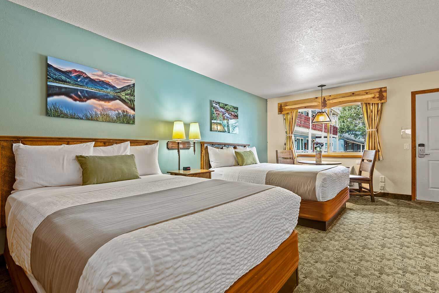 Room with two queen beds, green pillows, dining set next to the window and artwork of Colorado landscapes